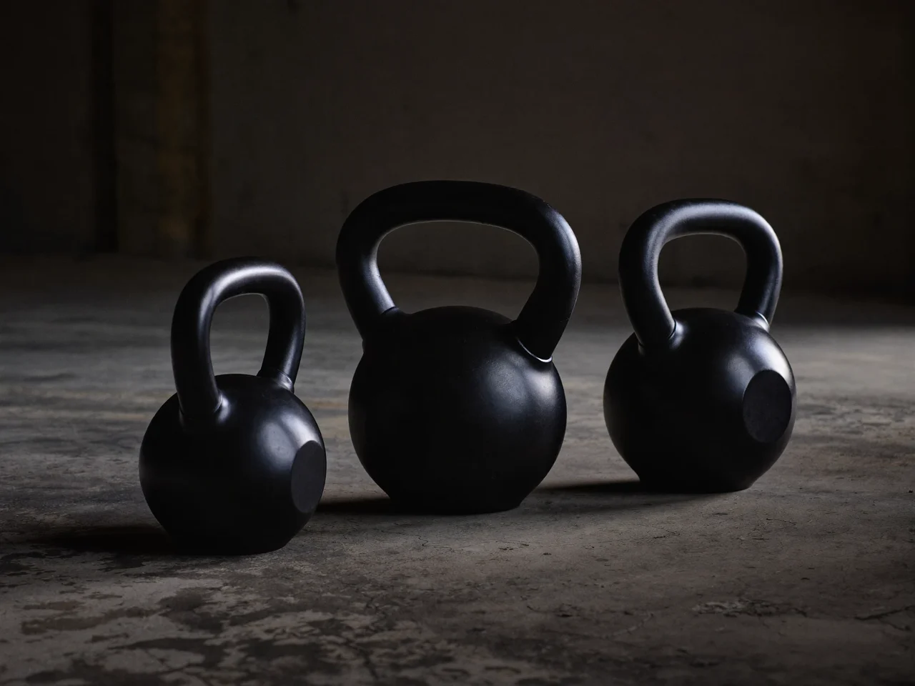 Drei Gusseisen-Kettlebells in abgestuften Grössen auf rohem Betonboden im Seitenlicht